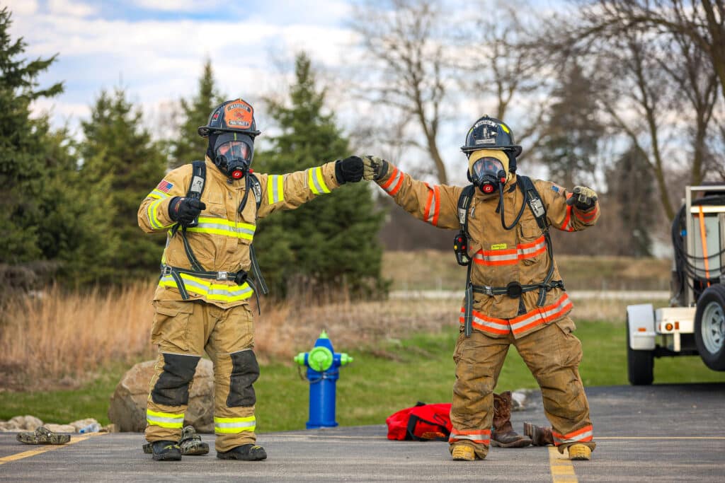 Two fire protection students in full gear fist-pumping in front of trees.