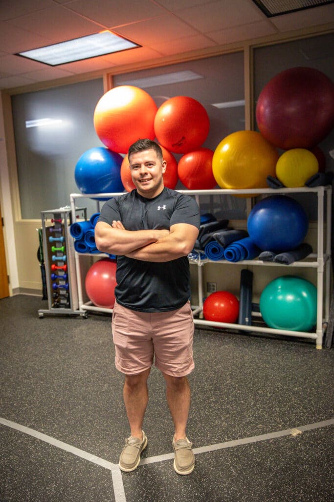 Smiling health and wellness student standing in front of rack of colorful gymnastics balls.