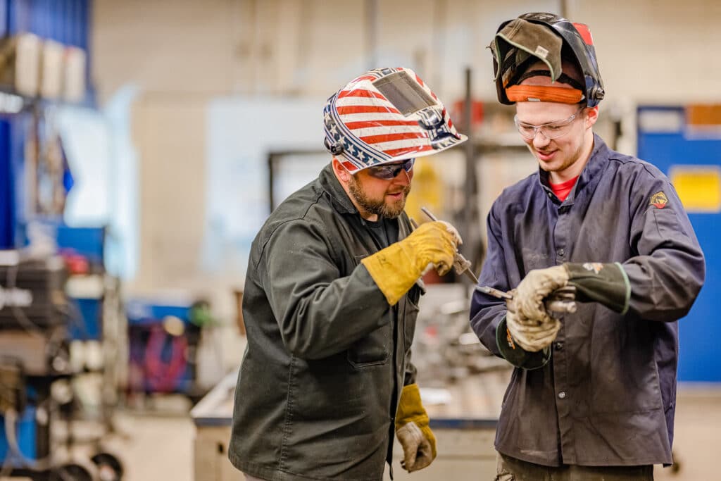 Teacher showing student in welding outfit a piece of metal.