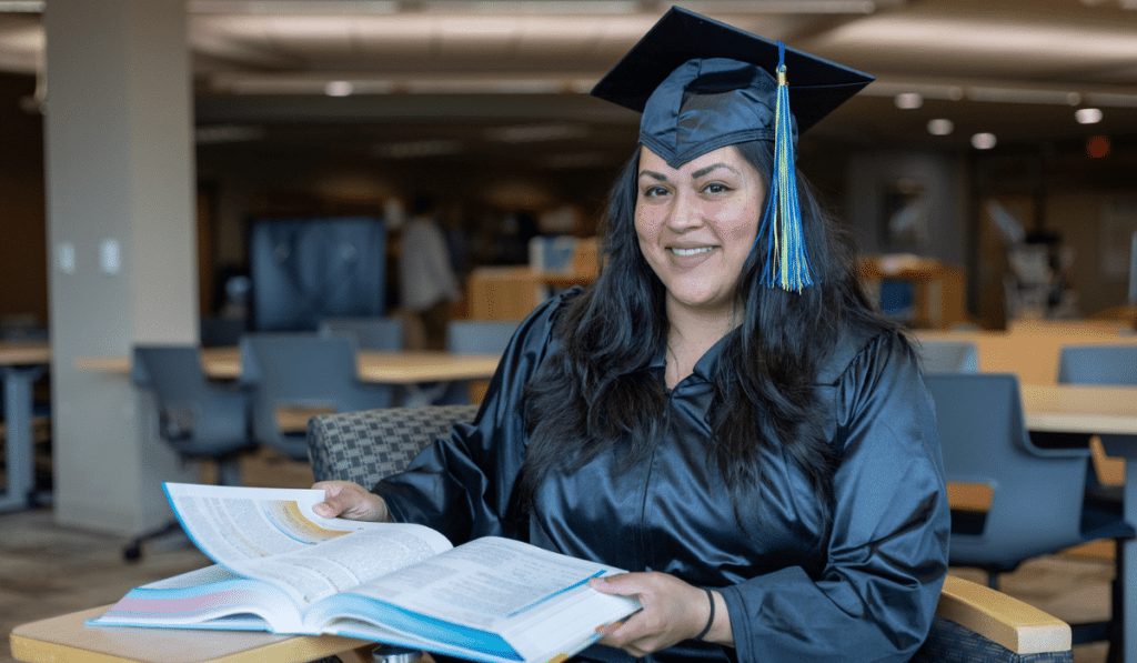 Moraine Park student Delilah Hernandez sitting at a table flipping through a text book while proudly wearing her graduation gown.